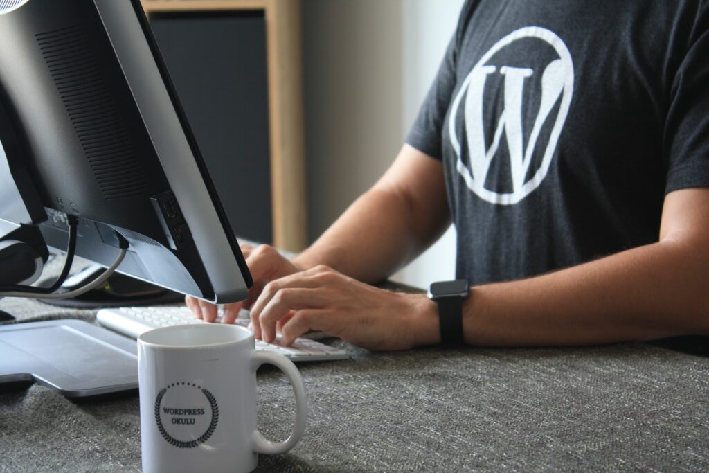 Someone typing at a computer wearing a T-shirt with the WordPress logo on it and a mug on the desk with WordPress written on it