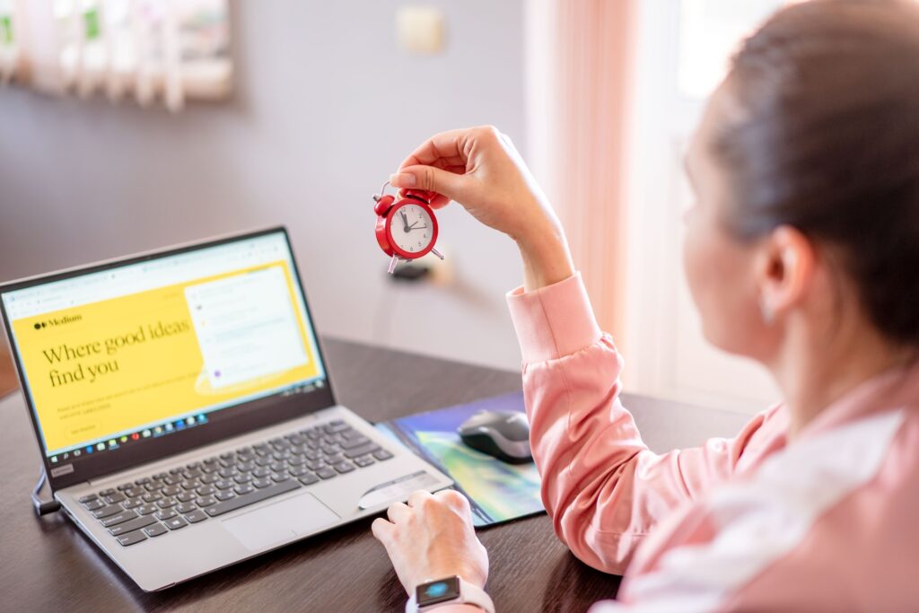 A lady sitting in front of her laptop holding a tiny alarm clock in her right hand