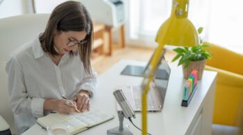 A lady sitting at her desk writing in a book with a laptop and microphone in front of her