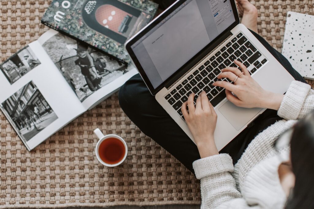 A lady typing on her laptop keyboard with books on the floor around her and a mug of tea next to her