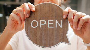 A woman holding a wooden speech bubble with the word OPEN written on it